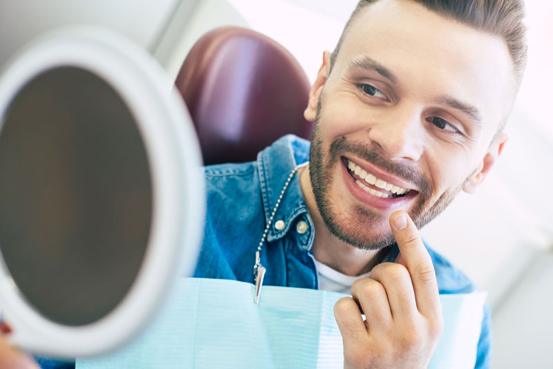 a dental patient smiling into a hand held mirror while pointing to his teeth because he just improved his smile with dental veneers.