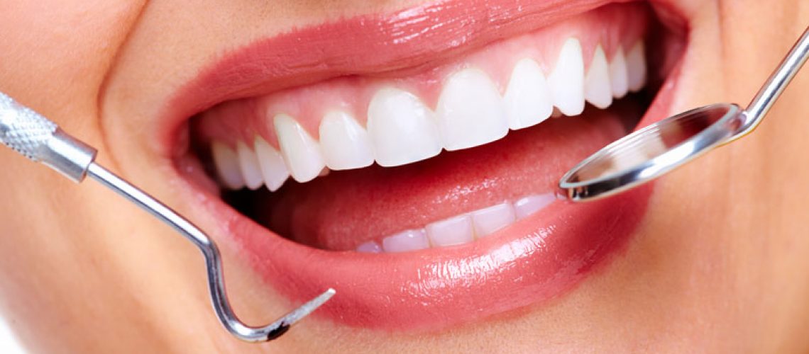 Close-up of a smiling mouth with white teeth being examined by dental tools, likely during a dental check-up or treatment.